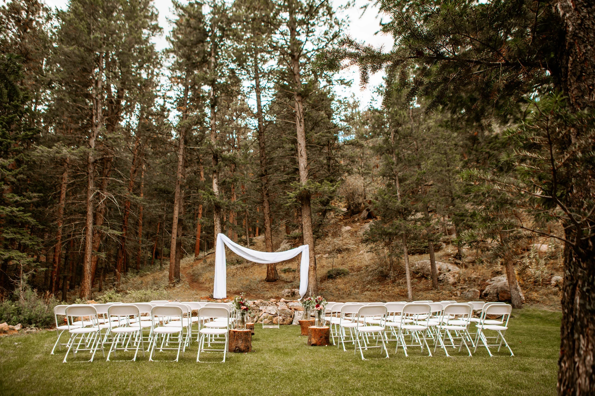 An outdoor wedding ceremony setup with white chairs, a draped arch, and floral decorations in a wooded area.